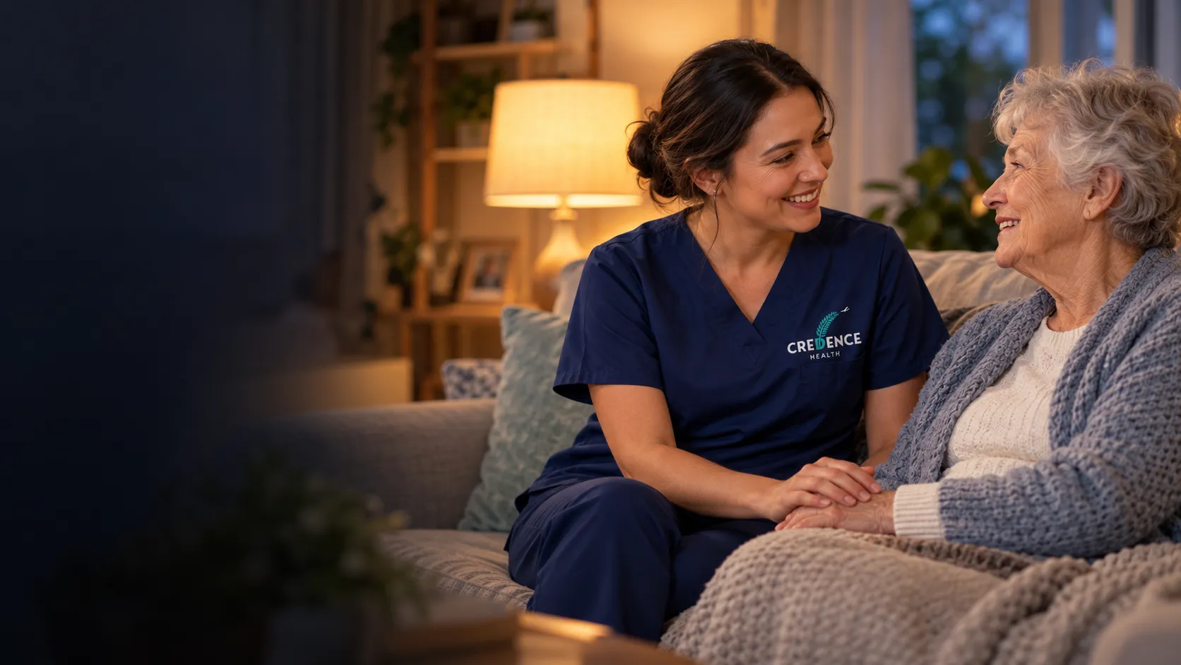 A nurse in a navy Credence Health uniform sits with an older woman in her living room, holding her hand as they talk